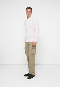 Camisa de botones rosa claro de manga larga combinada con pantalones cargo color camel que cuentan con múltiples bolsillos y zapatos negros.