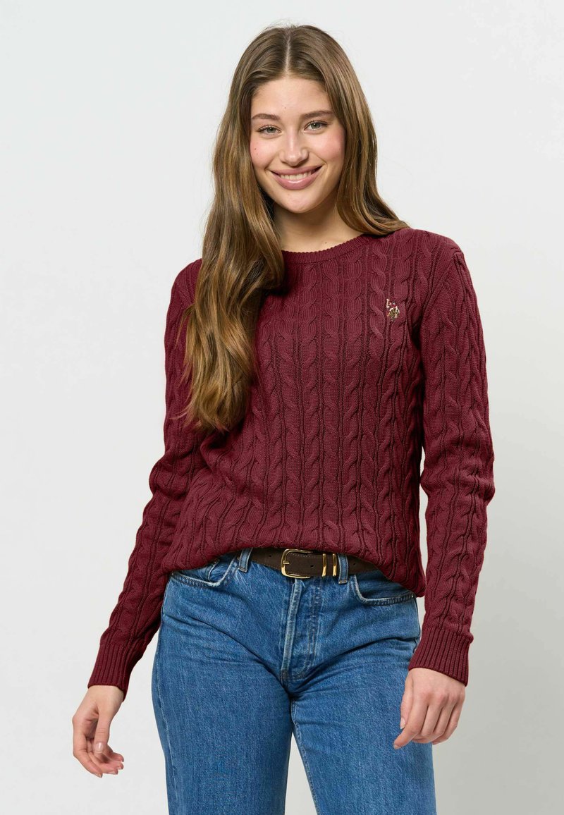 Jeune femme aux longs cheveux bruns sourit, portant un pull torsadé bordeaux et un jean bleu avec une ceinture marron sur un fond uni.