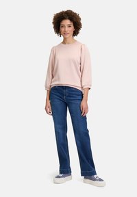 Ljusrosa sweatshirt med puffärmar, kombinerad med mörkblå raka jeans och sneakers i lila toner, står framför en neutral bakgrund.