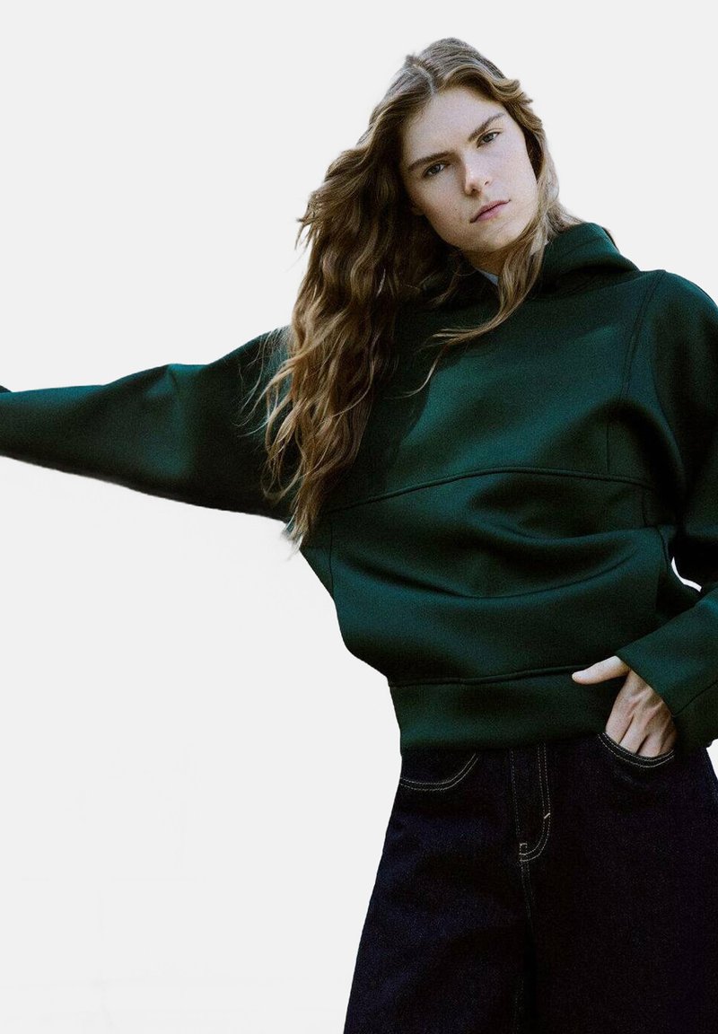 Sudadera oversized verde oscuro con un acabado texturizado, que cuenta con un bolsillo frontal y capucha con cordón, combinada con jeans oscuros de cintura alta.