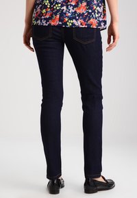 Tmavě modré džíny s přiléhavým střihem, charakteristickými zadními kapsami a zlatým stehem, spárované s černými botami. Květinová blůza je částečně viditelná.