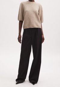 Beige Kurzarmpullover aus weichem, flauschigem Material, kombiniert mit lockeren schwarzen Hosen und spitzen schwarzen Absätzen. Schlichte, entspannte Silhouette.