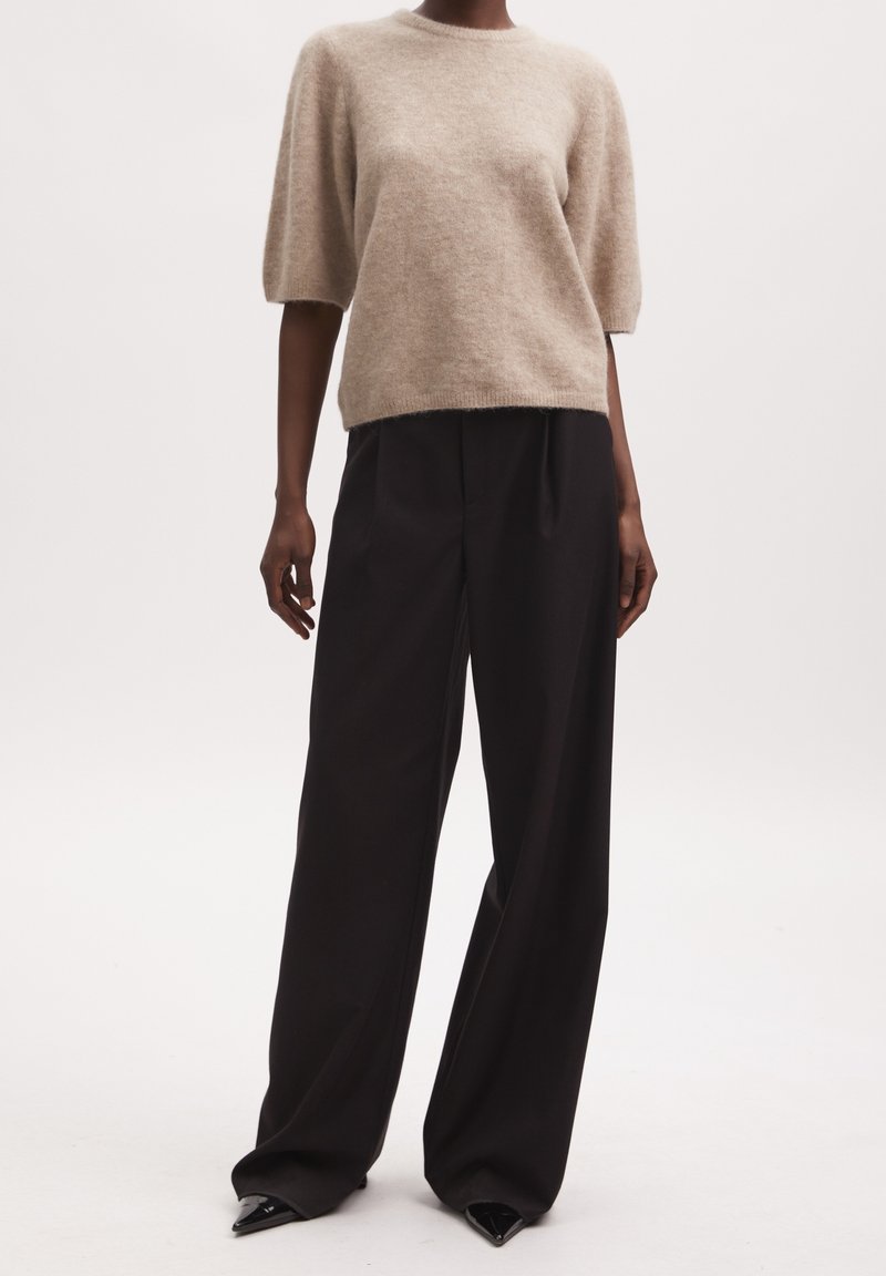 Beige Kurzarmpullover aus weichem, flauschigem Material, kombiniert mit lockeren schwarzen Hosen und spitzen schwarzen Absätzen. Schlichte, entspannte Silhouette.