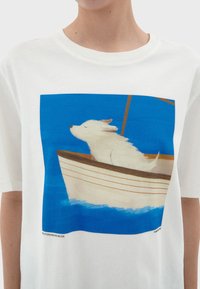 Witte t-shirt met een kleurrafische afbeelding van een hond op een boot, met een blauwe achtergrond en lichtbruine accenten langs de rand van de boot.