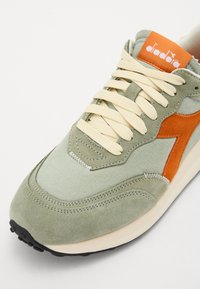 Green and orange athletic shoe featuring a combination of suede and canvas, round toe, and beige laces. Includes a visible logo on the tongue.