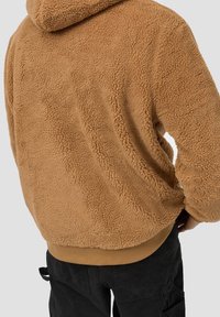 Ruskea fleece-huppari, jossa on teksturettu, pörröinen pinta, huppu ja ribbireunus. Yhdistetty mustiin housuihin, joissa on cargo-taskut.