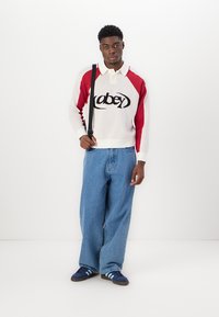 Vitt stickat tees med röda ärmar och svart "obey"-logotyp, parat med löst sittande blå denimjeans och marinblå skor med blå ränder.