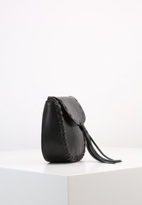 Czarna skórzana torba na ramię z dekoracyjnym przeszyciem wzdłuż krawędzi, klapką na zapięcie oraz długim detalem w postaci frędzla. Kompaktowy, zaokrąglony kształt.