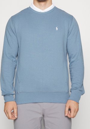 Mann trägt ein hellblaues Sweatshirt mit Rundhalsausschnitt über einem weißen Hemd mit Kragen und hellgrauen Hosen, steht mit locker an den Seiten hängenden Händen.