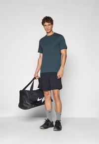 Türkiisi kaelusega t-särk, mustad lühikesed püksid, hallid sokid ja mustad kingad. Mudel hoiab suurt musta Nike reisikotti valge logo detailiga.
