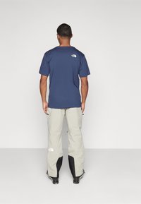 Navyblauw shirt met korte mouwen met logo op de bovenrug, gecombineerd met lichtgrijze broek met zwarte accenten en ritsen bij de zakken.