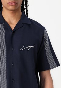 Camisa de manga corta de color navy con diseño de botones, que presenta un acento blanco estampado en un lado y un logotipo distintivo bordado en el pecho.