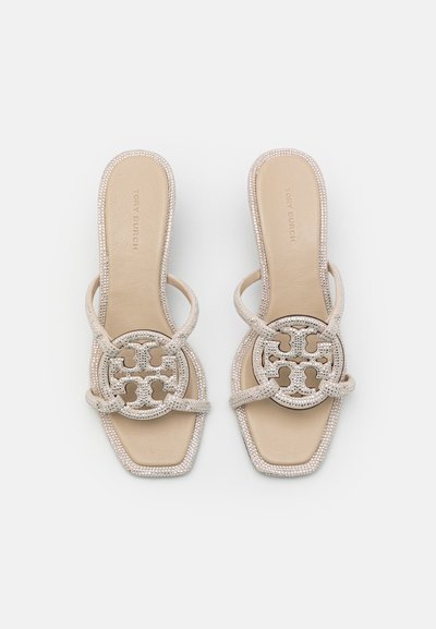 Sandales à enfiler en beige avec une semelle texturée. Possède un grand logo découpé décoratif avec des accents en strass et un design à bout carré.