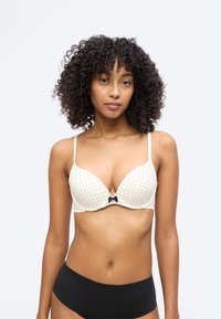 Νέα γυναίκα με σγουρά μαλλιά, φορούσα ένα λευκό σουτιέν με πουά σχέδιο και μαύρο φιόγκο, καθώς και μονόχρωμο μαύρο εσώρουχο με ψηλή μέση, στέκεται μπροστά σε ένα απλό φόντο.