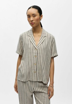 Vrouw draagt een beige kort mouw knoophemd en bijpassende broek met verticale zwarte strepen, staand tegen een effen witte achtergrond.