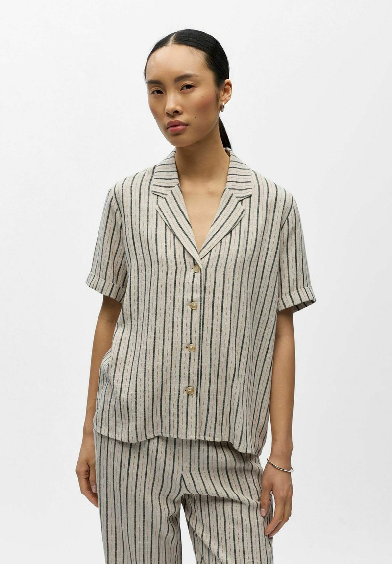 Femme portant une chemise beige à manches courtes boutonnée et un pantalon assorti avec des rayures verticales noires, debout devant un fond blanc uni.