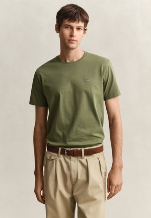 Jeune homme debout portant un t-shirt à manches courtes vert olive rentré dans un pantalon beige plissé avec une ceinture en cuir marron.