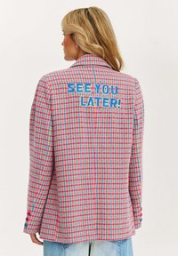 Žena s blonďatými vlasy, která má na sobě růžovo-modrý kostkovaný blazer s nápisem "UVIDÍME SE POZDĚJI!" v modrém textu na zádech, doplněný o světle modré džíny.