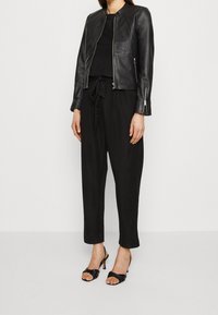 Chaqueta de cuero negra con cuello redondo, cierre de cremallera y mangas largas, llevada sobre una blusa negra y pantalones negros holgados, complementada con tacones.