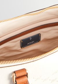 Sac à main beige ouvert avec une fermeture éclair dorée, bordure en cuir marron et une étiquette noire à l'intérieur portant la mention "JOOP!", fixée avec des rivets dorés.