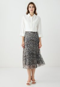 Weißes Hemd mit aufgerollten Ärmeln, kombiniert mit einem plissierten, knielangen Rock mit braunem Schlangenmuster. Weiße Sandalen mit offenen Zehen.