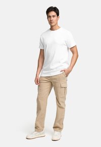 Vit t-shirt med kort ärm, beigefärgade cargo-byxor med sidofickor, bärs med vita sneakers. Modellen står självförtroendefullt mot en enfärgad bakgrund.