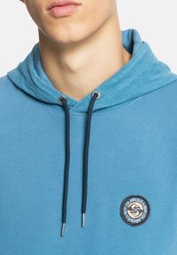 Modrá mikina s kapucí vyrobená z měkké látky, s tkaničkou na stažení kapuce. Na přední straně je zaoblená nášivka s logem a kontrastní textura na kapuci.