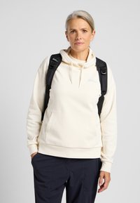 Cream-colored hoodie with a kangaroo pocket and drawstring hood, worn with black backpack straps, paired with navy pants.