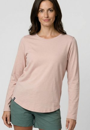 Frau mit schulterlangem braunem Haar, die ein hellrosa langärmeliges Shirt und grüne Shorts trägt, vor einem schlichten Hintergrund stehend.