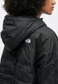 Czarna puchowa kurtka o strukturalnym wykończeniu, z kapturem i białym logo po lewej stronie klatki piersiowej. Posiada przeszyte kieszenie i regulowane paski.