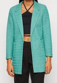 Türkiser Blazer mit Fischgrätenmuster, mit Reverskragen, zwei vorderen Taschen und lockerer Passform, kombiniert mit einem schwarzen Crop-Top und einer Hose.