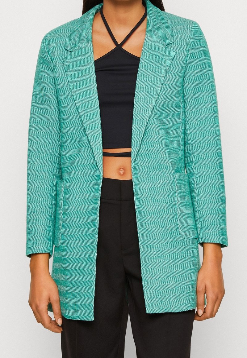 Türkiser Blazer mit Fischgrätenmuster, mit Reverskragen, zwei vorderen Taschen und lockerer Passform, kombiniert mit einem schwarzen Crop-Top und einer Hose.