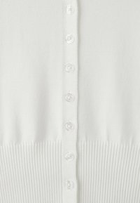 Gros plan sur un cardigan blanc avec une rangée de sept petits boutons blancs et une texture en maille côtelée au bord inférieur.