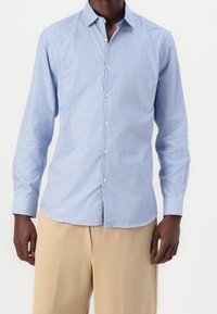 Hombre vestido con una camisa de botones de manga larga de cuadros en azul claro ajustada y pantalones beiges, de pie contra un fondo blanco.