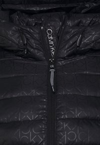 Black puffer jacket with Calvin Klein branding, featuring a zip front closure, a quilted design, and textured fabric with a subtle logo pattern.