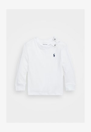 Balts garām piedurknēm krekls no kokvilnas ar apaļu kakla izgriezumu, kuram ir divi sudraba spiedpogas plecos un mazs tumši zils logo uz krūtīm.