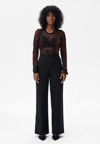 Vrouw met krullend haar draagt een doorschijnende zwarte top met bloemmotief en lange mouwen, een zwarte high-waisted broek met wijde pijpen, en zwarte puntige schoenen, staand rechtop.