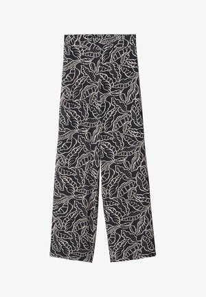 Femme portant un haut noir en maille à manches longues et un pantalon noir à jambes larges avec imprimé feuilles blanches, mains dans les poches, debout sur un sol gris.
