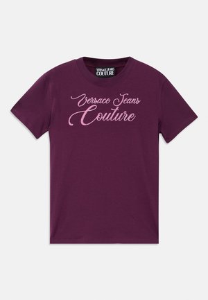 Vijolična bombažna majica s kratkimi rokavi, ki vsebuje bleščeč roza napis "Versace Jeans Couture" v kurzivni pisavi na sprednjem delu.