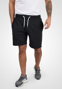 Shorts de sport noirs avec cordon de serrage, dotés d'une texture en coton lisse et de deux poches latérales, associés à des baskets grises et à un t-shirt blanc.