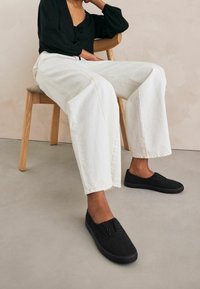 Schwarze, strukturierte Slippers mit fünf Ösen, kombiniert mit loser weißer Denim-Hose, sitzend auf einem Holzstuhl vor einem neutralen Hintergrund.
