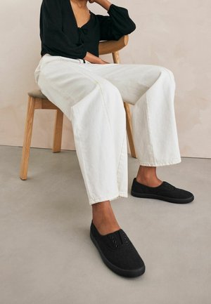 Zapatos negros de texturizados sin cordones con cinco ojales, combinados con pantalones de mezclilla blancos holgados, sentados en una silla de madera contra un fondo neutral.
