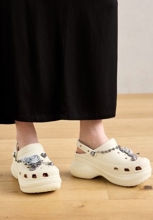 Pieds portant des sabots compensés écru décorés de breloques argentées, debout sur un sol en bois avec un pantalon large noir visible.