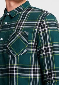Vihreä ja musta ruudullinen flanellipaita, jossa on nappikaulus, rintatasku ja valkoisia sekä keltaisia yksityiskohtia kuviossa.