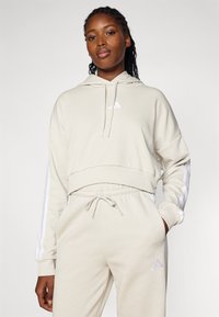 Light beige cropped hoodie with drawstring and white Adidas logo. Paired with matching tapered joggers featuring white side stripes.