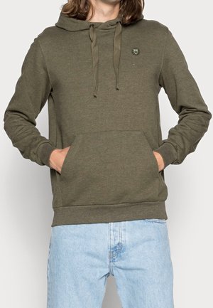 Person iført olivengrøn hættetrøje med frontlomme og emblem på brystet, kombineret med lyseblå jeans, hænderne i lommerne.