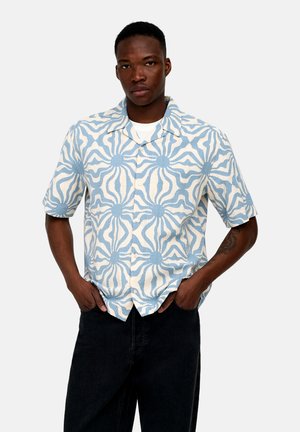 Férfi, rövid ujjú, gombos ingben, kék és fehér absztrakt nap mintával, fekete nadrággal, kezét a zsebében tartva.