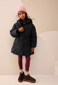 Manteau matelassé noir avec capuche, bonnet côtelé rose, leggings bordeaux et chaussures marron à motif léopard. Debout devant un fond clair.
