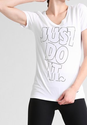 Vrouw draagt een witte V-hals T-shirt met de tekst "JUST DO IT." in grote, omlijnde letters, gecombineerd met een zwarte broek, en heeft één hand omhoog geheven.