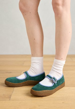 Green suede platform shoes with blue trim, featuring a silver buckle strap. Worn with white ankle socks on a wooden floor.
